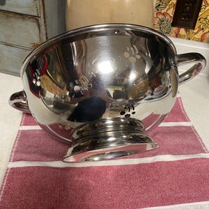 May include: A silver colander with a wide, round bowl and two handles. The colander has a textured surface with small holes throughout the bowl. The colander is sitting on a red and white striped kitchen towel.
