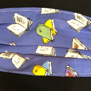 May include: A blue fabric face mask with a pattern of white and colored books. The mask has a white stitched seam around the edges.