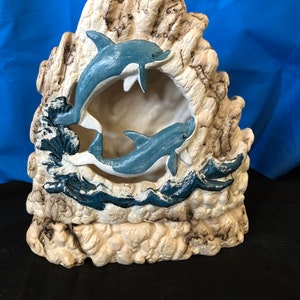 Puede incluir: Una figurita de cerámica de dos delfines saltando a través de una ola, sobre un fondo de formación rocosa beige. Los delfines están pintados en tonos de azul y blanco.