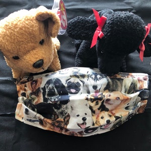 May include: A black face mask with a white background and a pattern of various dog breeds. The mask has black elastic straps. Two plush dog toys are in the background, one brown and one black.