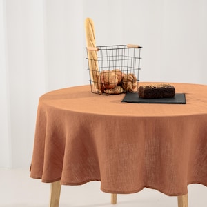 May include: A round table covered with a solid terracotta-colored linen tablecloth. On the table sits a black wire basket containing a baguette and several round bread rolls, alongside a dark rectangular slate board holding a loaf of seeded bread.