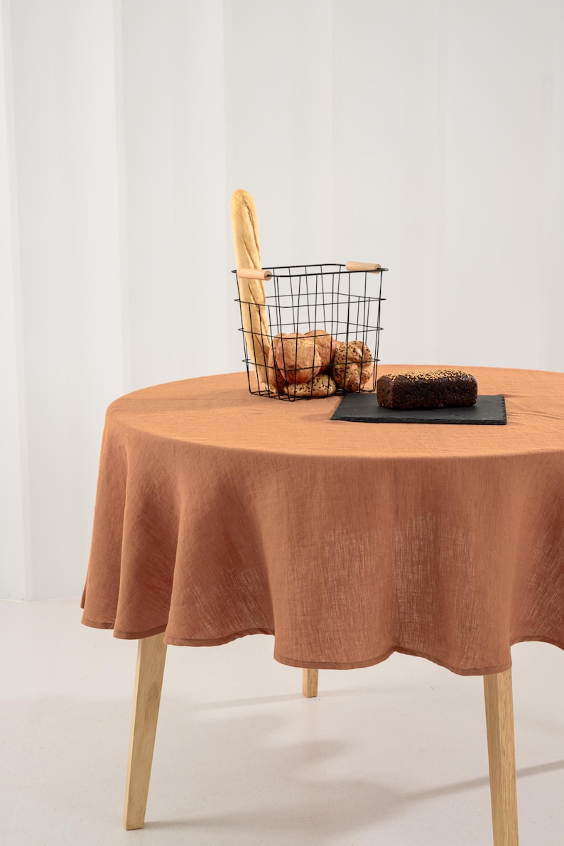 May include: A round table covered with a solid terracotta-colored linen tablecloth. On the table sits a black wire basket containing a baguette and several round bread rolls, alongside a dark rectangular slate board holding a loaf of seeded bread.