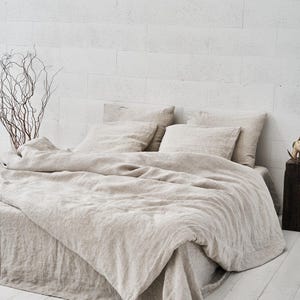 May include: A beige linen duvet cover and pillowcases are arranged on a bed with a white brick wall background. The bedding is made of natural linen fabric and has a relaxed, casual look.