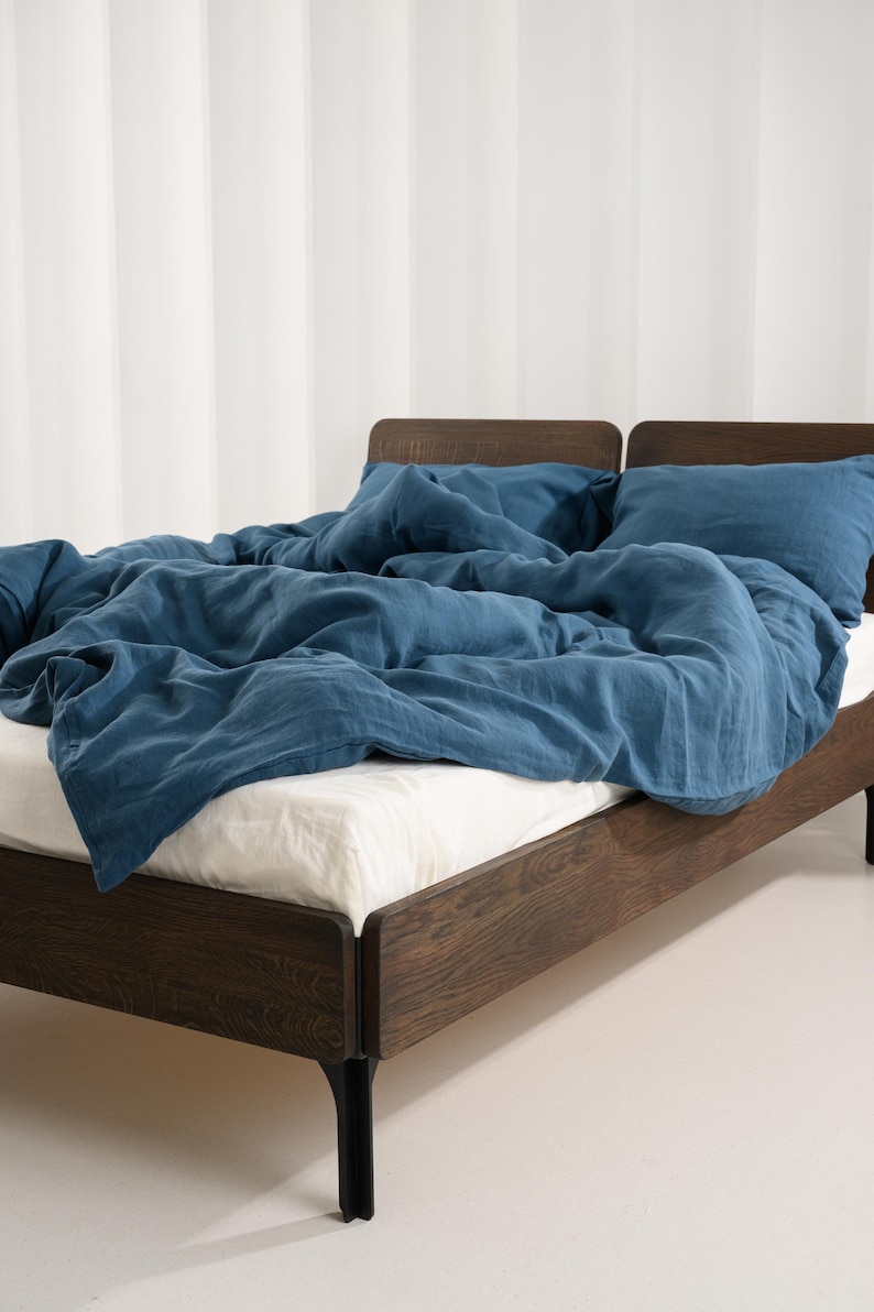 May include: A bed with a dark wood frame and black legs. The bed is covered with a blue comforter and pillows. The sheets are white. The background is a white wall with vertical stripes.