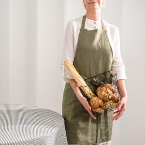 May include: An olive green linen apron worn over a white long-sleeved shirt, paired with a black wire basket containing a long baguette and several round, seeded bread rolls. The apron features a simple neck strap and waist ties.