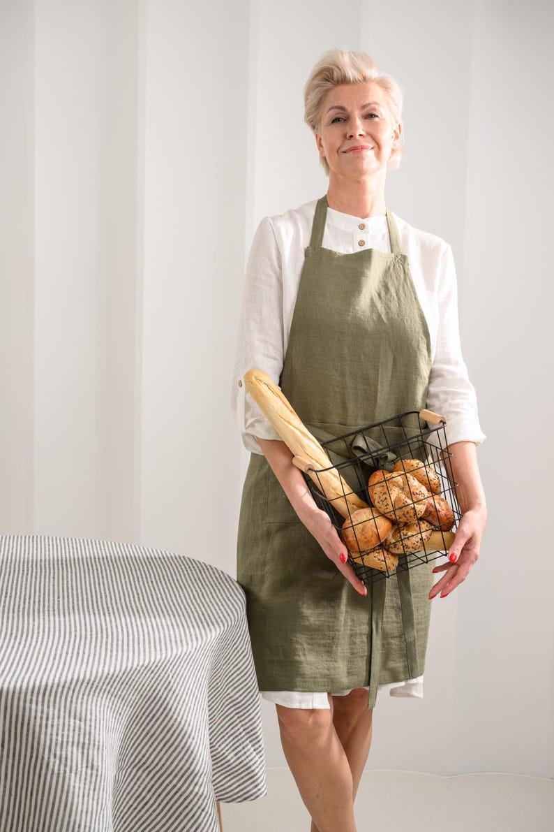 May include: An olive green linen apron worn over a white long-sleeved shirt, paired with a black wire basket containing a long baguette and several round, seeded bread rolls. The apron features a simple neck strap and waist ties.