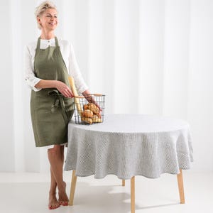 May include: An olive green linen apron with a tie waist and two front pockets, worn over a white long-sleeved shirt, paired with a round table covered in a white and grey striped tablecloth and a black wire basket filled with assorted bread.