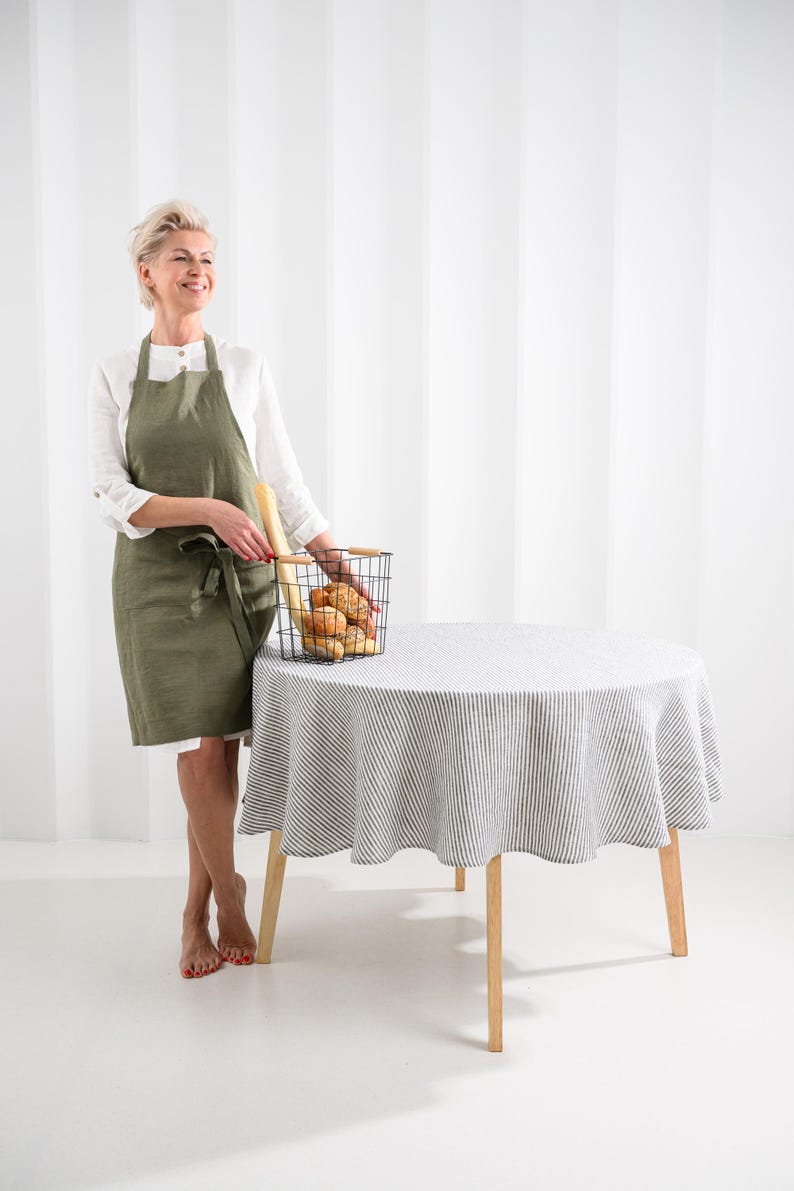 May include: An olive green linen apron with a tie waist and two front pockets, worn over a white long-sleeved shirt, paired with a round table covered in a white and grey striped tablecloth and a black wire basket filled with assorted bread.