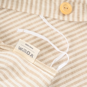 May include: Close-up of a beige and white striped fabric with a button and a tag that reads "100% linen". White drawstrings are attached to the fabric. The fabric has a natural, textured appearance.