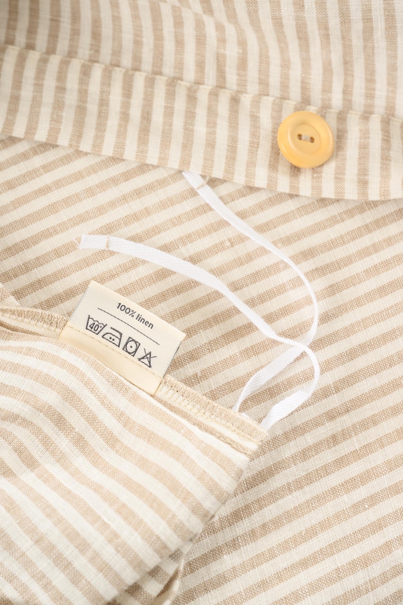 May include: Close-up of a beige and white striped fabric with a button and a tag that reads "100% linen". White drawstrings are attached to the fabric. The fabric has a natural, textured appearance.