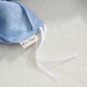 May include: Close-up of a light blue linen face mask with white drawstrings. The mask has a tag that reads "Linen Hygge". The mask is laying on a light beige linen surface.