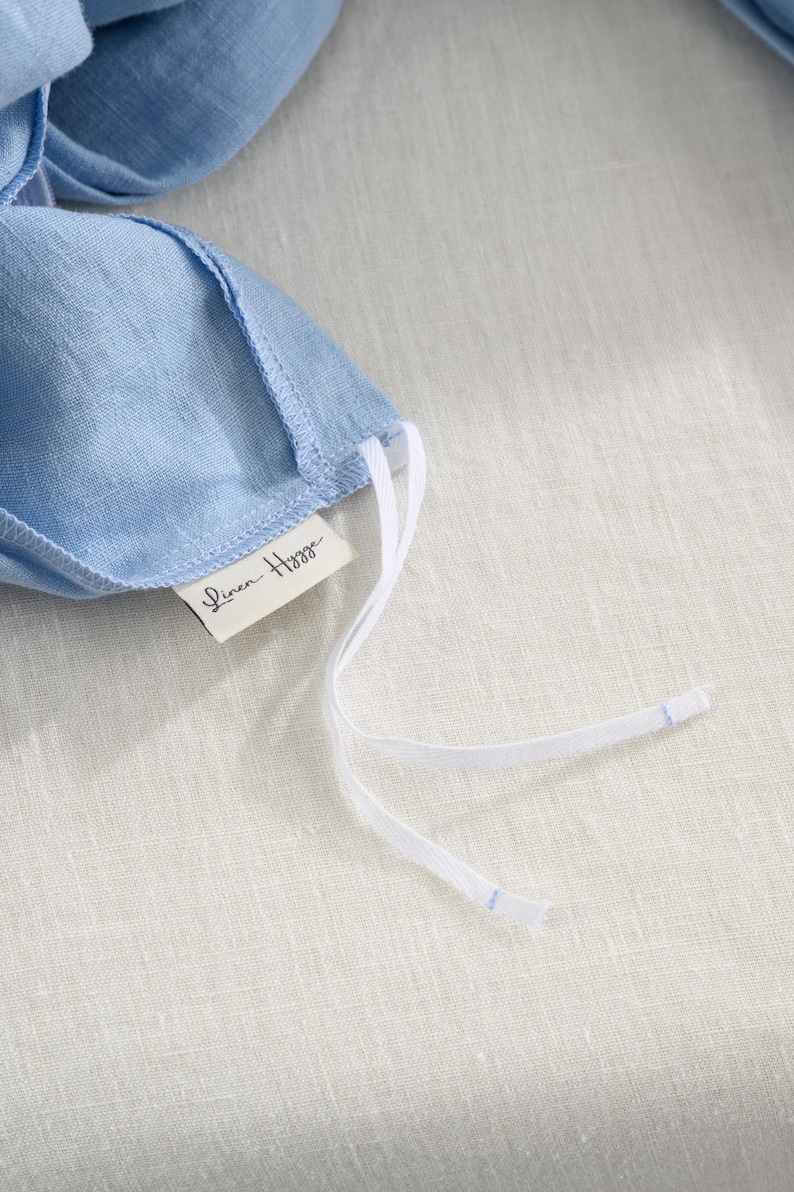May include: Close-up of a light blue linen face mask with white drawstrings. The mask has a tag that reads "Linen Hygge". The mask is laying on a light beige linen surface.