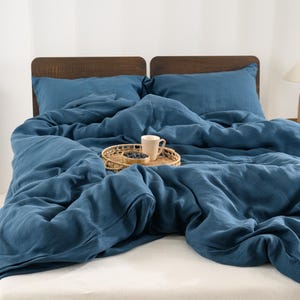 May include: A bed with a blue linen duvet cover and matching pillowcases. A small woven tray holds a mug and a bowl of berries. The headboard is made of dark wood. A lamp sits on a side table.