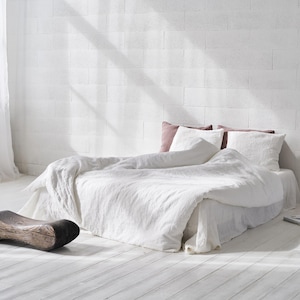 May include: A bed with white linen bedding, two white pillows, and one mauve pillow. A curved, dark brown wooden stool sits on the floor near the bed. A tall, thin brown vase containing dried grass stalks stands next to the bed on the floor.