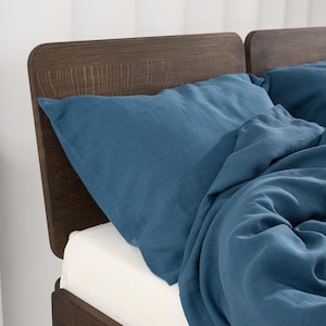 May include: A bed with a dark brown wooden headboard and a blue linen pillow and duvet. The bed has a white sheet. The headboard has a curved top and a textured wood grain.