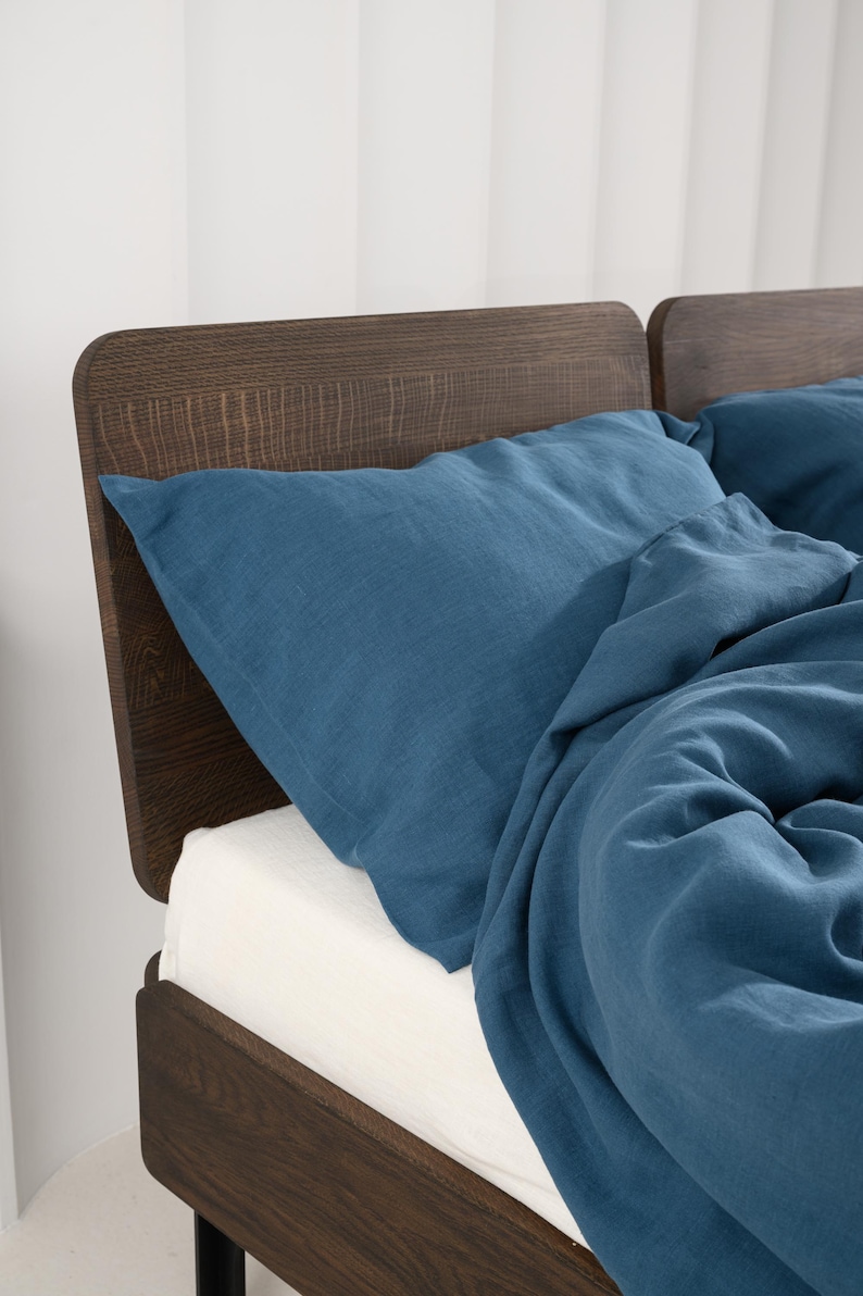 May include: A bed with a dark brown wooden headboard and a blue linen pillow and duvet. The bed has a white sheet. The headboard has a curved top and a textured wood grain.