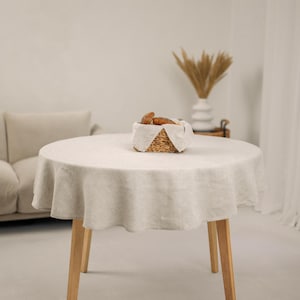 May include: A round, cream-coloured linen tablecloth on a wooden table. A woven basket containing bread sits on the table. The room is bright and minimalist, with a sofa and a decorative vase in the background.