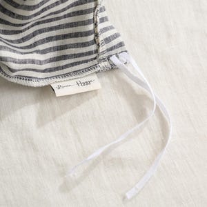 May include: Close-up of a striped fabric sun hat in gray and white. The hat has white drawstrings and a small tag that reads "Linen Hygge". The hat is made of a soft, textured fabric.