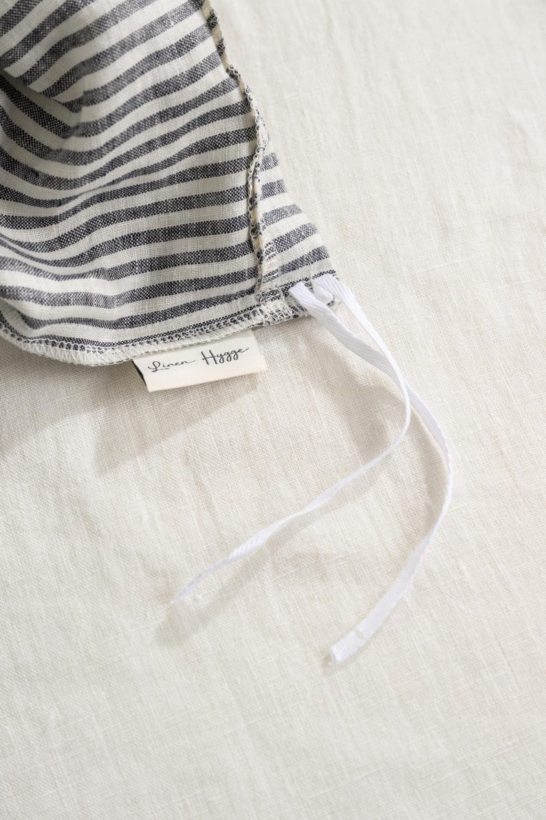May include: Close-up of a striped fabric sun hat in gray and white. The hat has white drawstrings and a small tag that reads "Linen Hygge". The hat is made of a soft, textured fabric.