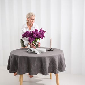 May include: A round dark gray linen tablecloth draped over a wooden table. On the table sits a glass vase filled with purple lilac flowers, a stack of white plates, a folded black and white checkered napkin, and a tall thin beige candle.