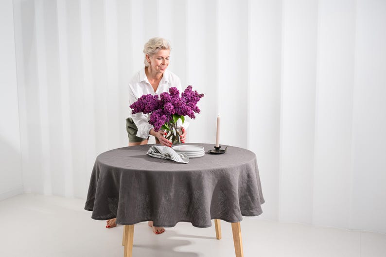 May include: A round dark gray linen tablecloth draped over a wooden table. On the table sits a glass vase filled with purple lilac flowers, a stack of white plates, a folded black and white checkered napkin, and a tall thin beige candle.