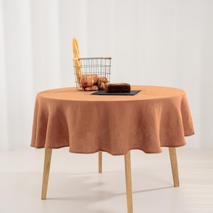 May include: A round, terracotta-colored linen tablecloth draped over a circular wooden table with four light-colored legs. A black wire basket containing a baguette and bread rolls sits on the table next to a dark rectangular slate serving board.