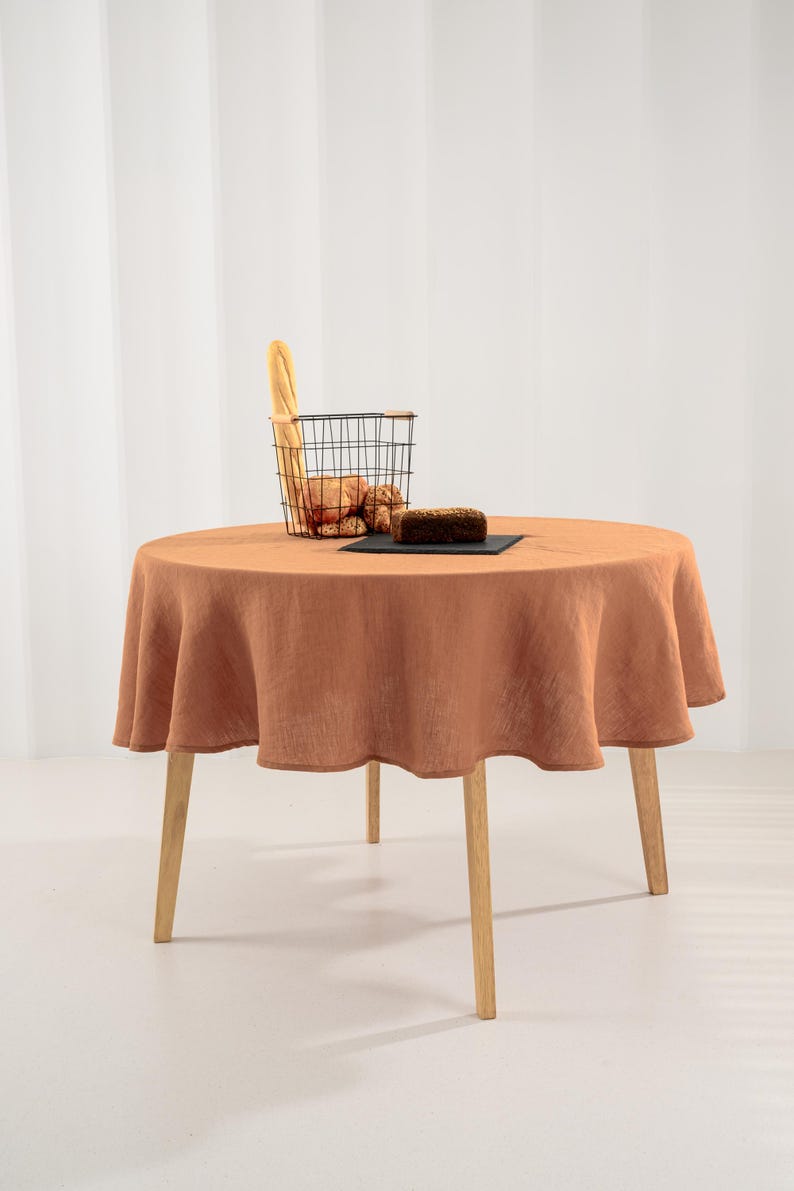May include: A round, terracotta-colored linen tablecloth draped over a circular wooden table with four light-colored legs. A black wire basket containing a baguette and bread rolls sits on the table next to a dark rectangular slate serving board.