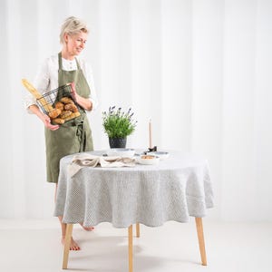 May include: A round table covered with a grey and white striped linen tablecloth, featuring a potted lavender plant, a single taper candle, and small ceramic bowls. An olive green apron is worn over a white shirt, holding a black wire basket of bread.