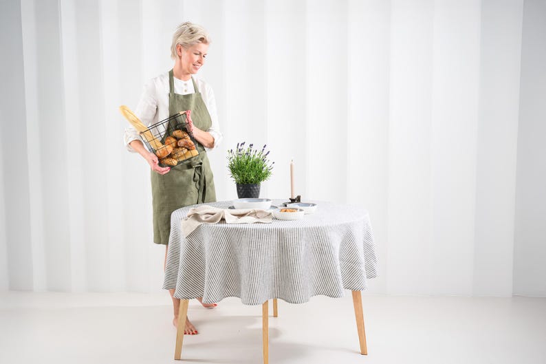 May include: A round table covered with a grey and white striped linen tablecloth, featuring a potted lavender plant, a single taper candle, and small ceramic bowls. An olive green apron is worn over a white shirt, holding a black wire basket of bread.