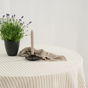 May include: A round table covered with a beige and white striped tablecloth. On the table sits a black textured pot containing a lavender plant, a beige taper candle in a black metal holder, and a folded beige linen napkin beside the candle.