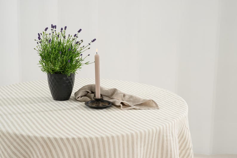 May include: A round table covered with a beige and white striped tablecloth. On the table sits a black textured pot containing a lavender plant, a beige taper candle in a black metal holder, and a folded beige linen napkin beside the candle.