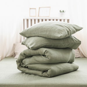 May include: A stack of sage green linen bedding, including a duvet cover and pillowcases, on a bed with white sheets.