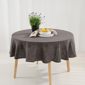 May include: A round table with light-colored wooden legs covered by a dark charcoal gray linen tablecloth. On the table sits a black pot containing a green lavender plant, a stack of white ceramic plates, and two yellow lemon halves.