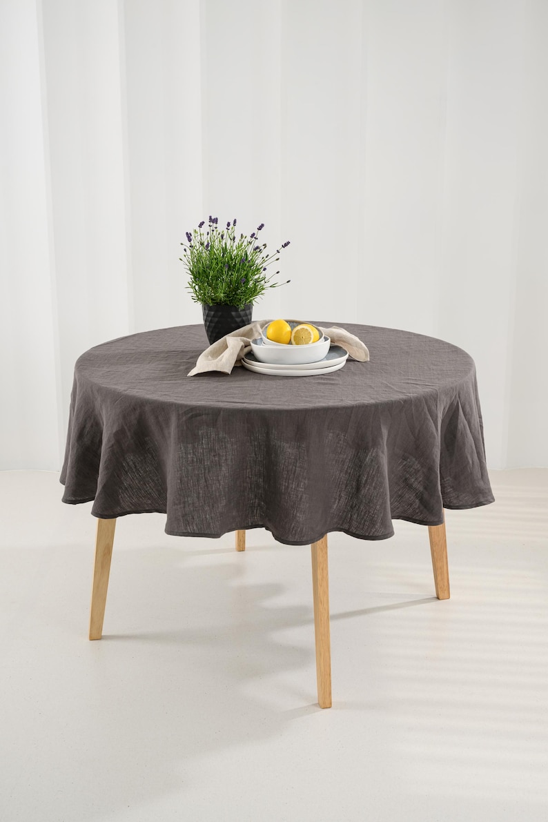 May include: A round table with light-colored wooden legs covered by a dark charcoal gray linen tablecloth. On the table sits a black pot containing a green lavender plant, a stack of white ceramic plates, and two yellow lemon halves.