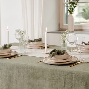 Può includere: Una tovaglia di lino verde con un runner bianco e tre coperti. Ogni coperto comprende una ciotola, un piatto, un tovagliolo, una forchetta, un coltello e un bicchiere di vino. Ci sono tre candele bianche sul tavolo.