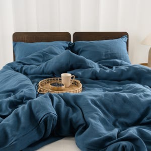 May include: A bed dressed with blue linen bedding, including a duvet and pillows. A woven tray holds a mug and a small bowl of berries. A lamp with a beige base and white shade sits on a small side table. The headboard is dark wood.