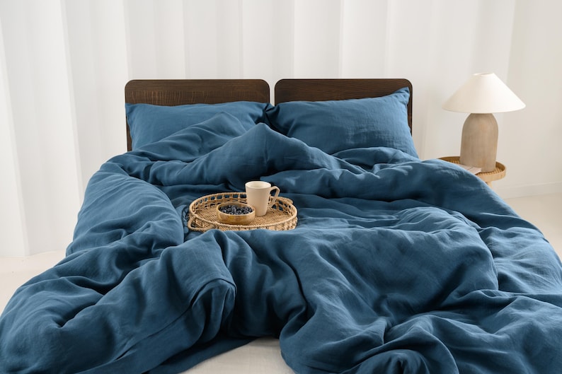 May include: A bed with blue bedding, including a duvet and pillows. A woven tray holds a mug and a small bowl of berries. A lamp with a beige base and white shade sits on a small side table. The headboard is made of dark wood.