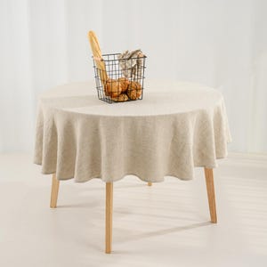May include: A round table covered with a beige linen tablecloth featuring a natural textured weave. On the table sits a black wire basket containing a long baguette and several round bread rolls wrapped in a light-colored checkered cloth napkin.