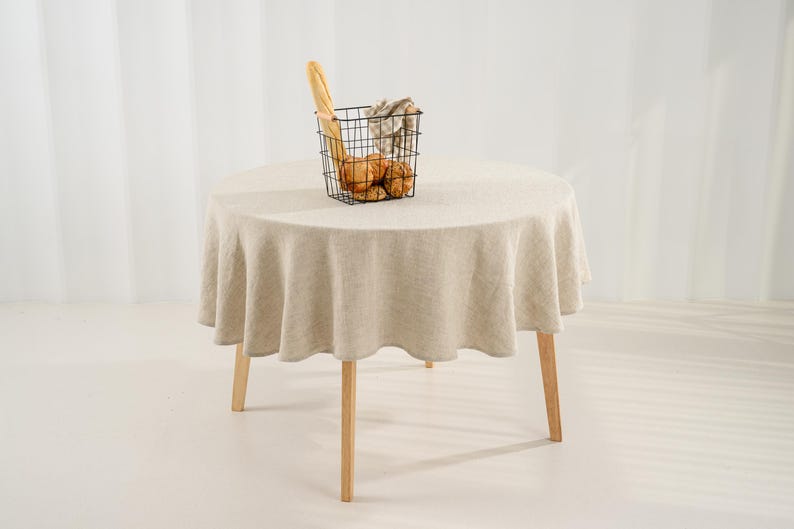 May include: A round table covered with a beige linen tablecloth featuring a natural textured weave. On the table sits a black wire basket containing a long baguette and several round bread rolls wrapped in a light-colored checkered cloth napkin.