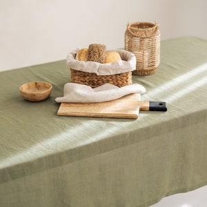 May include: A green linen tablecloth with a wooden cutting board, a woven basket filled with bread, a small wooden bowl, and a woven lantern.