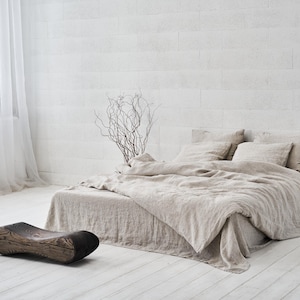 May include: A light beige linen bedding set with a natural wood stool in a minimalist bedroom setting. The bed is made with a linen duvet cover and several pillows. The room has white walls and a sheer white curtain.