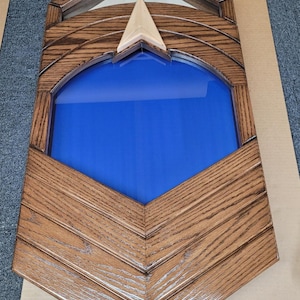 May include: Wooden display case with a blue interior. The case has a shield shape with a light-colored wooden arrow-like accent. The frame is made of stained wood with a chevron pattern.