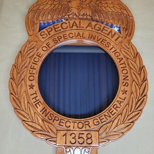 May include: A wooden plaque with carved details, featuring the words "SPECIAL AGENT" and "OFFICE OF SPECIAL INVESTIGATIONS". An eagle is carved at the top. The plaque has a circular opening with a blue background and the number "1358".