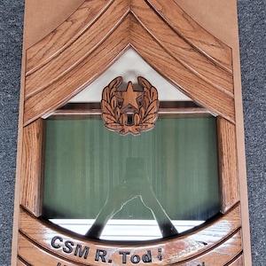 Puede incluir: Caja de sombra militar de madera con forma de galón. La caja presenta un emblema de estrella y corona de laurel, una parte delantera de cristal y el texto grabado: "CSM R. Tod", "United States Army" y las fechas "14 de julio de 1994 - 31 de julio de 2024".