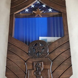 Op de afbeelding: Een donkerbruine houten display case in de vorm van een militair rangteken. Het heeft een gedeelte voor een vlag, een ster en een medisch embleem. De case heeft een blauwe achtergrond en is ontworpen om memorabilia in te bewaren.
