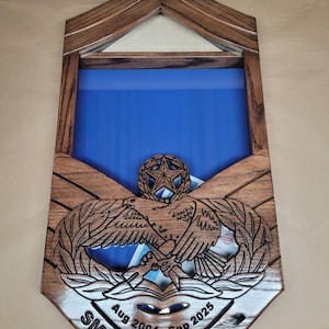 May include: Wooden display case with a blue background, featuring an eagle emblem and text that reads "SMSgt Chad Aug 2004 - Sep 2025." The case has a triangular top and a shield-like shape.