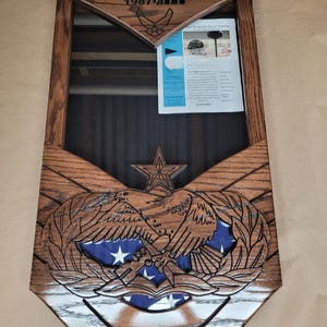 May include: A wooden display case with a dark brown finish, featuring an engraved eagle emblem and a star at the bottom. The top section is engraved with "TSgt Bobby Gannon 1987" and an Air Force logo. A folded American flag is visible inside.