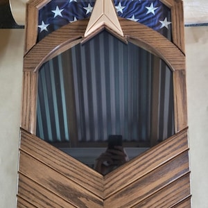 May include: A wooden flag display case with a blue fabric background featuring white stars. The case has a geometric design with a central mirror and a decorative wooden arrow. The frame is made of stained wood.