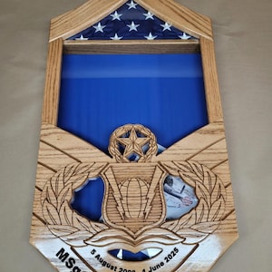 May include: Wooden display case with a blue interior, designed to hold a flag and other memorabilia. The case features a carved wooden frame, a section for a flag with white stars on a blue background, and a space for a military emblem. The name "MSgt Shawna R." is engraved.