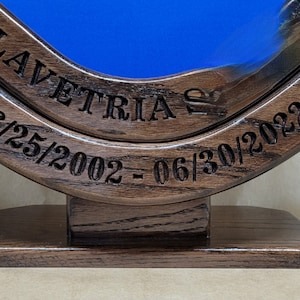 May include: A circular dark brown wooden frame with a blue center, featuring the engraved text ST LAVETRIA and the dates 06/25/2002 - 06/30/2022. The frame is mounted on a rectangular wooden base with a visible wood grain texture.